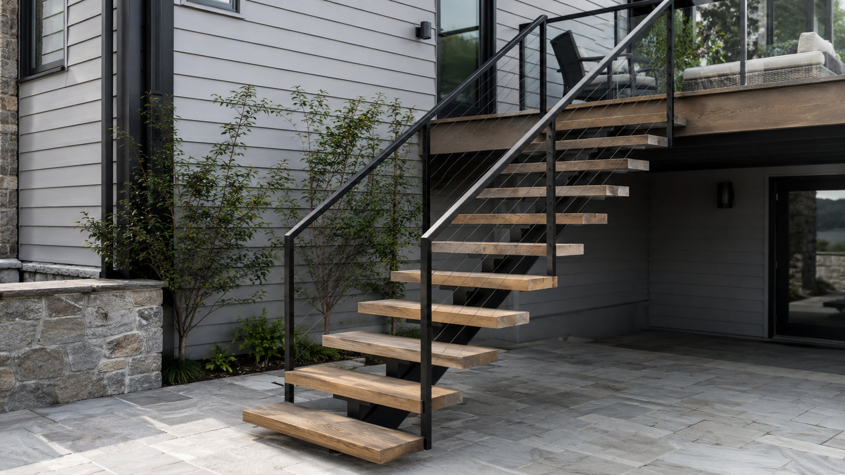 Exterior floating stairs with wood treads and cable railing connecting a modern home deck to a stone patio